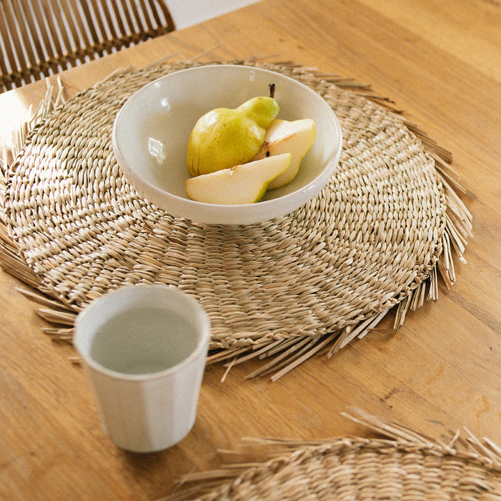 A half-slide look at a round seagrass woven placemat with a white plate above