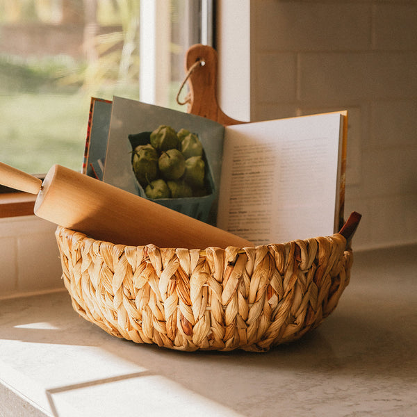 Artera Home round water hyacinth wicker storage basket with wooden handles storing a chopping board and a book